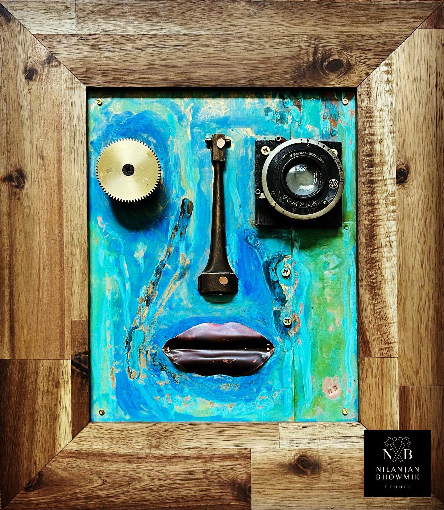 A wall-mounted art piece resembling a face, composed of old camera lens and clock parts, with a copper-colored lip and a brass base.